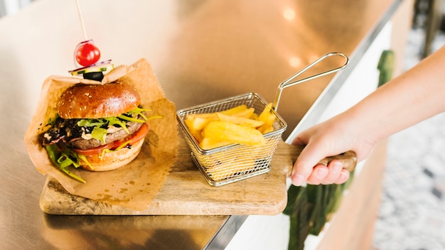 Mano de primer plano con tablero de madera con hamburguesa y papas fritas