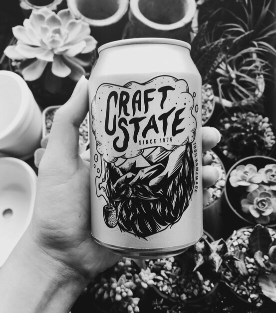 Mano que sostiene la lata de cerveza del estado del arte con el fondo de las plantas de interior