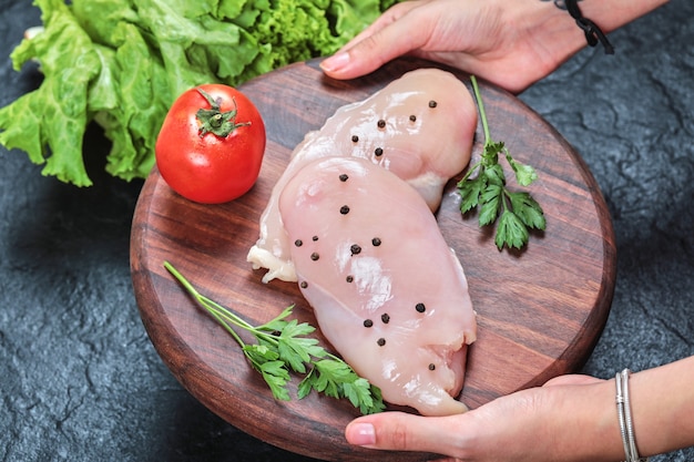 Mano que sostiene la placa de madera de filete de pollo crudo con verduras en la mesa oscura.