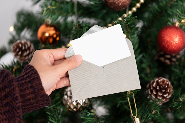 Mano que sostiene la tarjeta de felicitación de Navidad de la maqueta para el diseño de la invitación en el fondo del árbol de Navidad