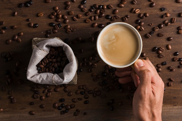 Mano que sostiene la taza con café cerca de saco con frijoles