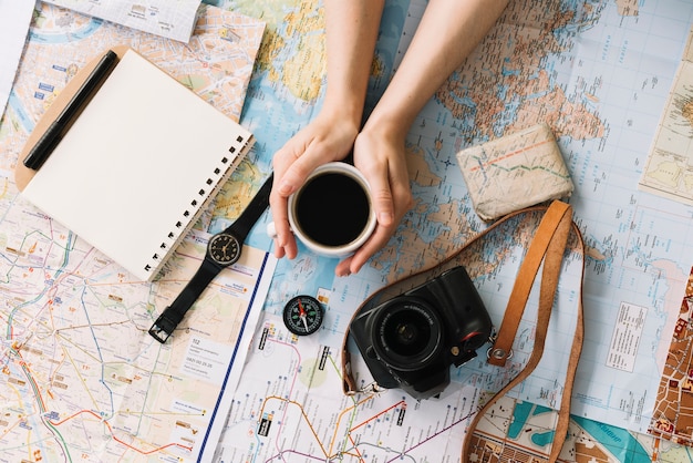 Mano que sostiene la taza de café en el mapa rodeado de cuaderno de espiral; Brújula; reloj de pulsera; cámara y correa