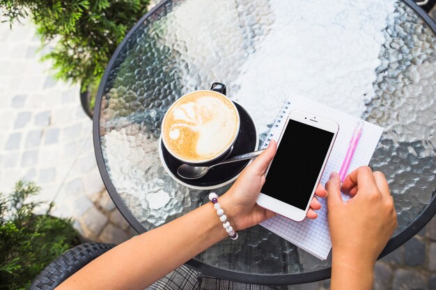 Mano que sostiene el teléfono móvil con taza de café en la mesa de cristal