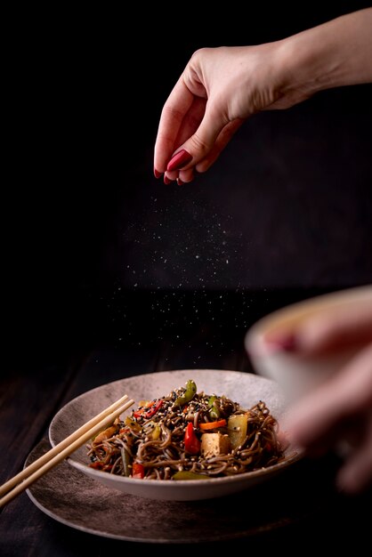 Mano rociando sal en un tazón de fideos con verduras