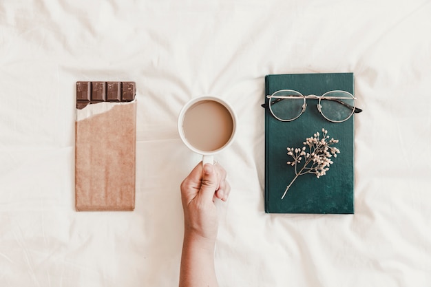 Foto gratuita mano sosteniendo bebida caliente cerca de libro y chocolate en sábana