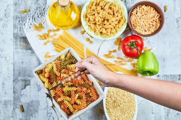 Mano sosteniendo una canasta de pasta fusilli cruda con pastas variadas y vegetales sobre la mesa de mármol.