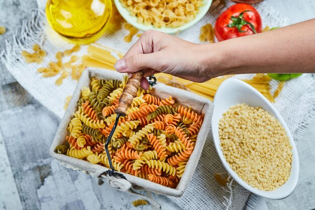 Mano sosteniendo una canasta de pasta fusilli cruda con una variedad de pastas y vegetales en la mesa de mármol.