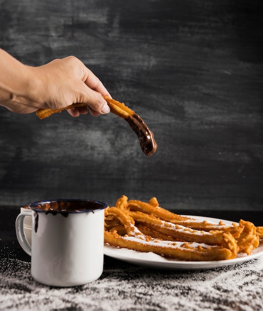 Mano sosteniendo un churros con chocolate y una taza