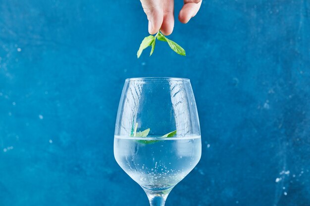 Mano sosteniendo menta encima de jugo en superficie azul