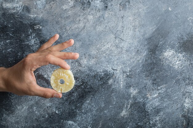 Mano sosteniendo una rodaja de piña seca sobre un fondo gris.