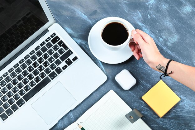 Mano sosteniendo una taza de té con laptop, notebook, lápiz y blocs de notas en el escritorio azul.