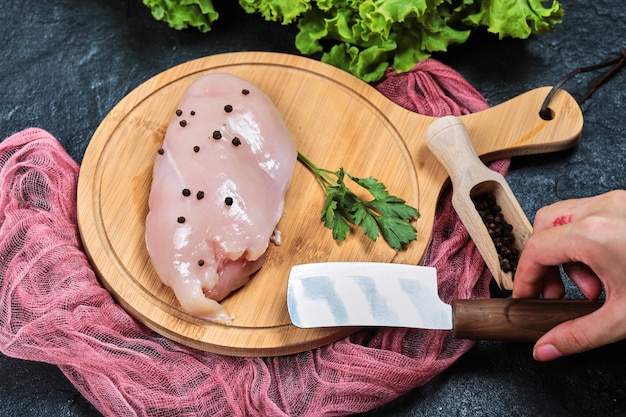 Mano sujetando un cuchillo y un trozo de filete de pollo crudo en un plato de madera con verduras frescas y mantel.