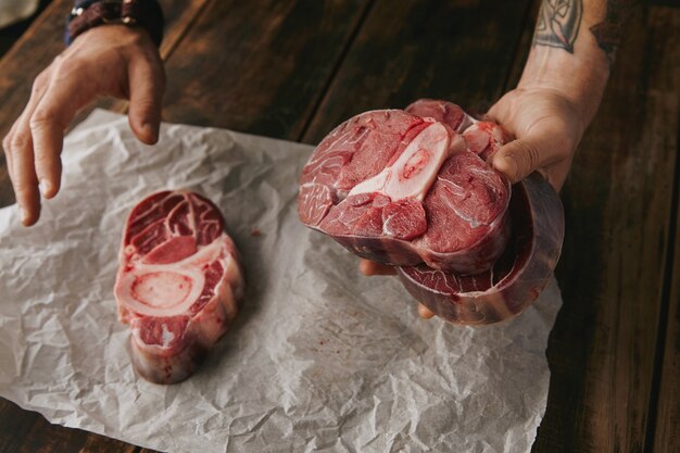 Mano tatuada ofrece un trozo de gran filete de carne cruda con hueso en la cámara, otros filetes en la mesa de madera vieja