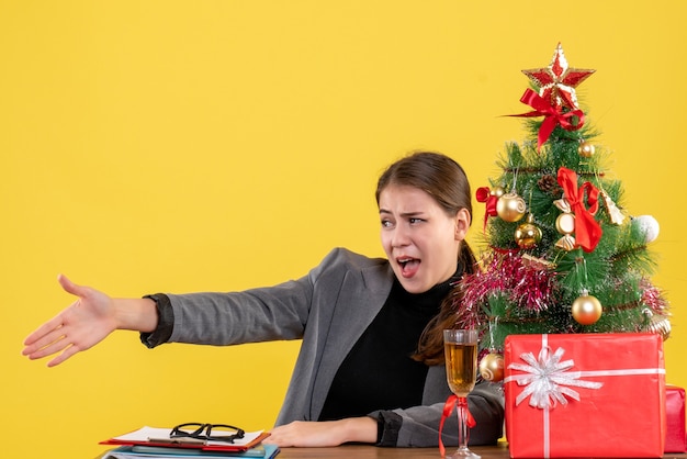 Mano de vista frontal dando chica pensativa sentada en el cóctel de árbol de Navidad y regalos de escritorio