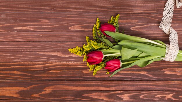 Manojo de flores de tulipán rojo y flores de vara de oro atadas con encaje sobre fondo de madera