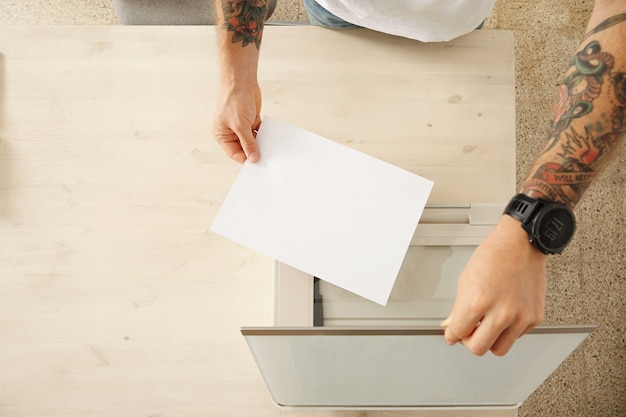 Las manos abren una bandeja de escáner y colocan una hoja de papel para escanear un documento en un dispositivo electrónico multifuncional doméstico, aislado en una mesa de madera blanca, vista superior