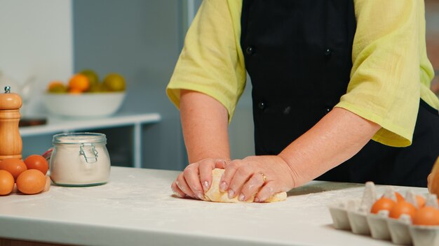 Manos de la abuela preparando galletas caseras en la cocina moderna amasando sobre la mesa. Panadero de ancianos jubilado con bonete mezclando ingredientes con harina de trigo para hornear pan y pasteles tradicionales