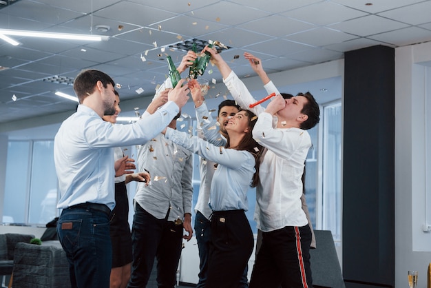 Foto gratuita manos arriba. foto del equipo joven en ropa clásica que celebra el éxito mientras sostiene bebidas en la moderna oficina bien iluminada