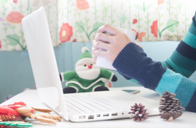 Manos celebración taza de café y el uso de ordenador portátil con la decoración de Navidad, Compras en línea