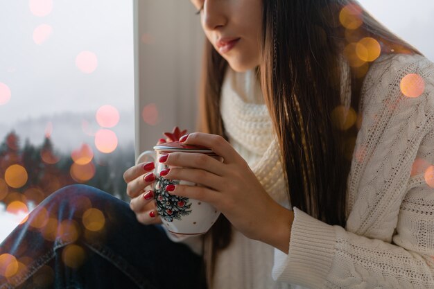 Manos cerca de mujer joven atractiva en elegante suéter de punto blanco sentado en casa en el alféizar de la ventana en Navidad sosteniendo la taza bebiendo té caliente, vista de fondo de bosque de invierno, luces bokeh