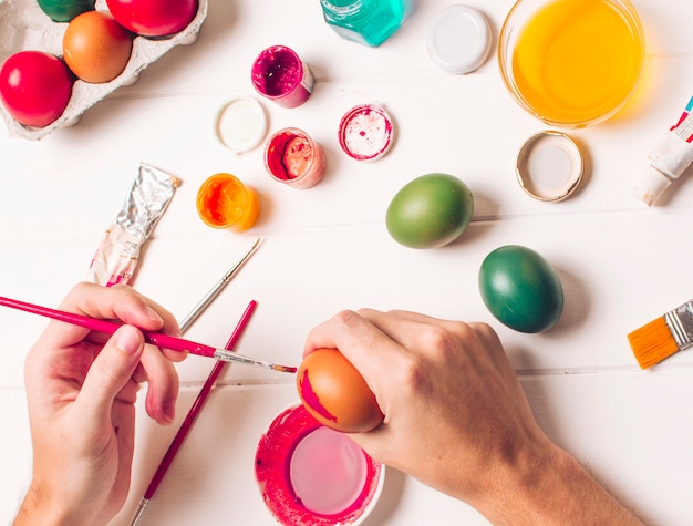 Manos para colorear huevos de Pascua cerca del recipiente, pinceles y tinte líquido en un tazón