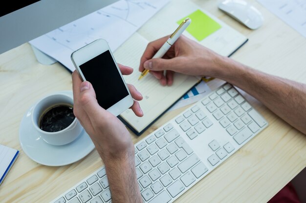 Las manos del diseñador gráfico masculino escribiendo en un diario y sostiene el teléfono móvil