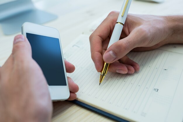 Las manos del diseñador gráfico masculino escribiendo en un diario y sostiene el teléfono móvil