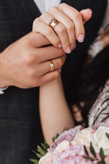 Manos de una esposa y un esposo con anillos de boda y compromiso y parte del ramo floral