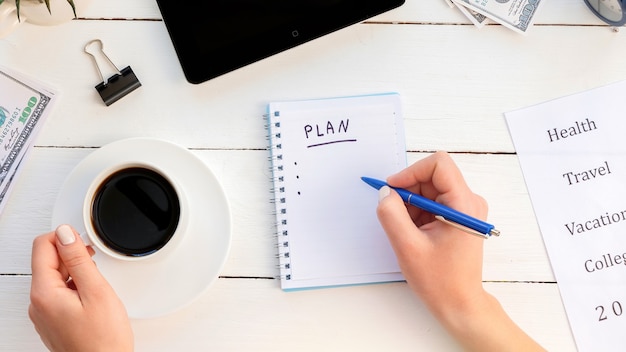 Manos femeninas escribiendo un plan de tareas en un bloc de notas, sosteniendo un café. Tableta, dinero. Fondo de madera