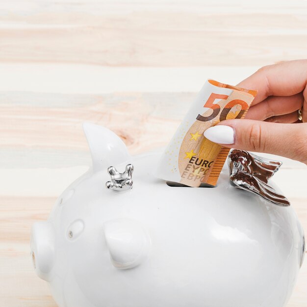 Manos femeninas que insertan cincuenta billetes de euros en el piggybank blanco