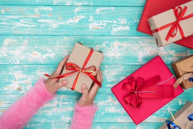 Manos femeninas sosteniendo una pequeña caja con un regalo entre decoraciones festivas de invierno en una vista superior de la mesa blanca. Composición plana para cumpleaños, navidad o boda.