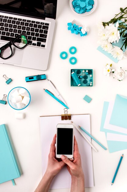 Las manos femeninas y el teléfono inteligente contra objetos de mujer de moda azul sobre blanco. Concepto de maqueta femenina