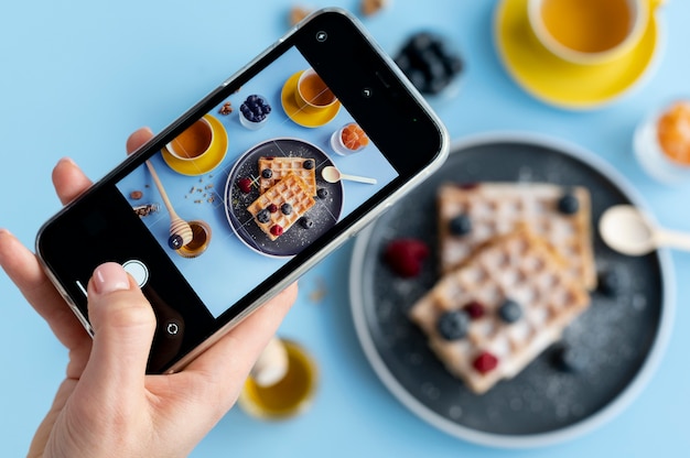 Manos femeninas tomando fotos de gofres con frutas y dos tazas de té