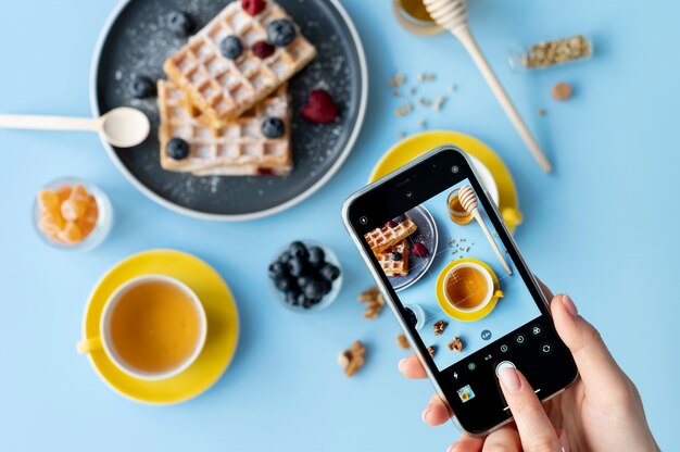 Manos femeninas tomando fotos de gofres con frutas y una taza de té