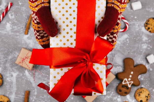 Las manos con guantes calientes sostienen la caja presente sobre la decoración de Navidad en el piso