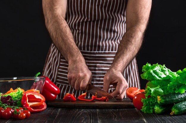 Manos de hombre cortando pimiento maduro en rodajas sobre una mesa. Marido en la cocina preparando una ensalada. sobre fondo negro