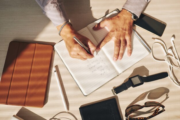 Manos del hombre escribiendo en diario y gadgets en la mesa