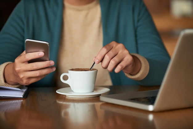 Manos del hombre irreconocible sentado en la cafetería, sosteniendo el teléfono inteligente y revolviendo el café