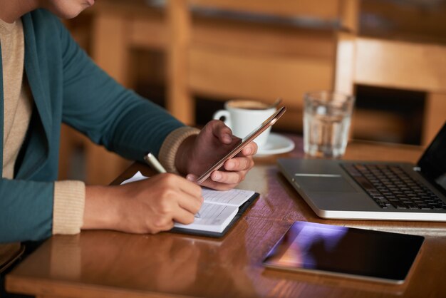 Manos de hombre irreconocible sentado a la mesa en la cafetería con gadgets y escribiendo en el cuaderno