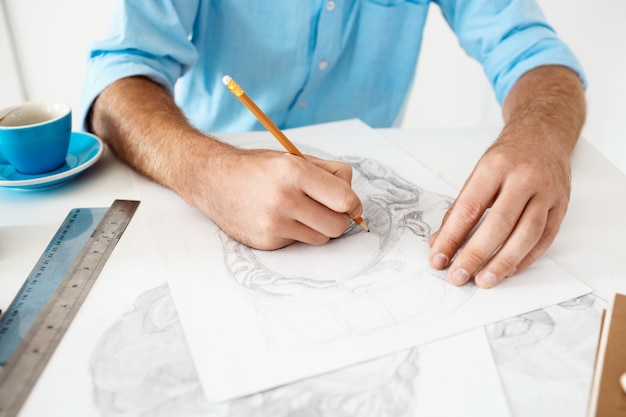 Manos del hombre de negocios joven que se sienta en la tabla con el retrato del dibujo de lápiz. Interior de oficina moderno blanco.