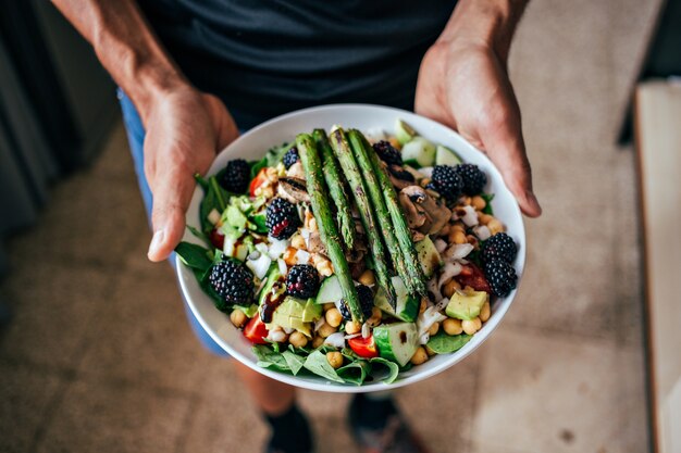 Manos de hombre sosteniendo un plato hondo grande lleno de ensalada vegetariana paleo saludable hecha de ingredientes biológicos orgánicos frescos, verduras y frutas, bayas y otras cosas nutricionales