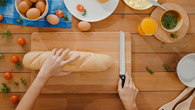 Las manos de la joven mujer asiática chef sostienen el cuchillo cortando pan integral sobre tabla de madera