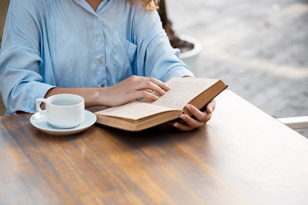 Manos de joven sentado en la mesa con libro y taza de café.