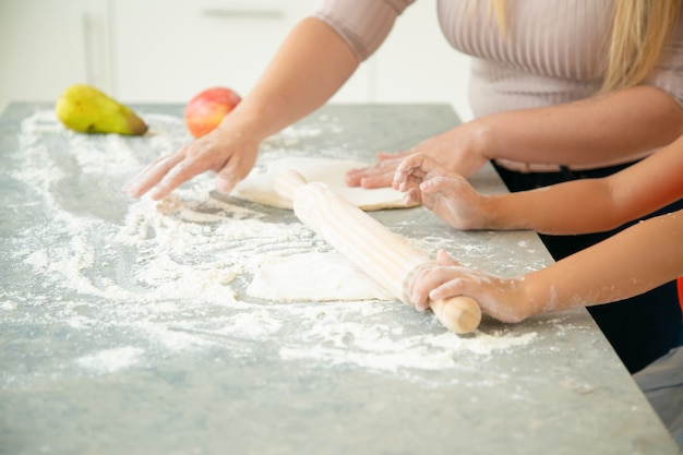 Manos de mamá e hija rodando masa sobre la mesa de la cocina. Niña y su madre horneando pan o pastel juntos. Primer plano, disparo recortado. Concepto de cocina familiar