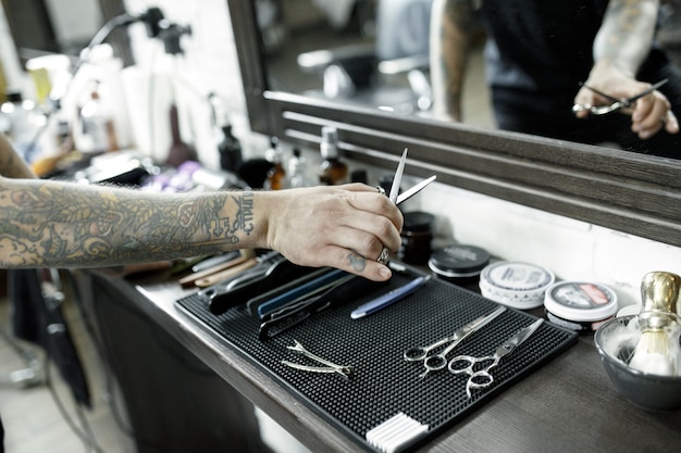 Las manos masculinas y herramientas para cortar la barba en la barbería. Herramientas vintage de peluquería. La mano del maestro tiene tatuaje con la palabra afeitar