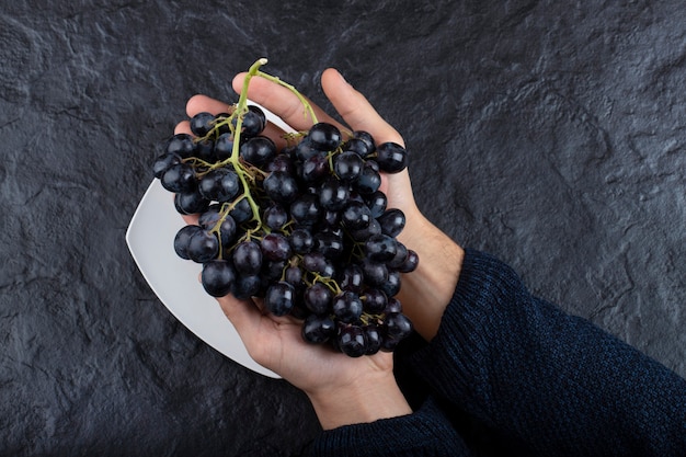 Manos masculinas sosteniendo racimo de uvas negras sobre fondo negro.