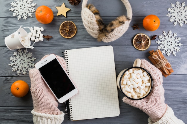 Manos en mitones con teléfono inteligente y taza con malvavisco cerca de cuaderno y copos de nieve de papel