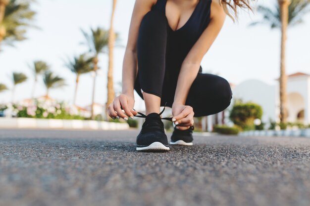 Manos de mujer bonita atado los cordones de las zapatillas de deporte en la calle con palmeras. Mañana soleada en ciudad tropical, entrenamiento, fitness, entrenamiento, motivación