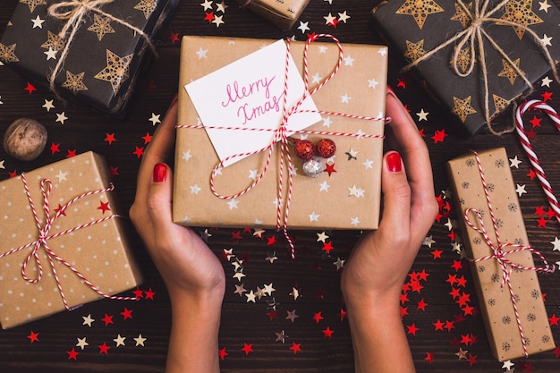 Manos de mujer con caja de regalo navideña con tarjeta postal feliz Navidad en mesa festiva decorada