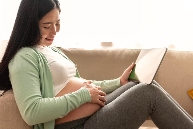 Las manos de una mujer embarazada asiática acarician el vientre y sostienen una pantalla de clave de croma verde en la tableta
